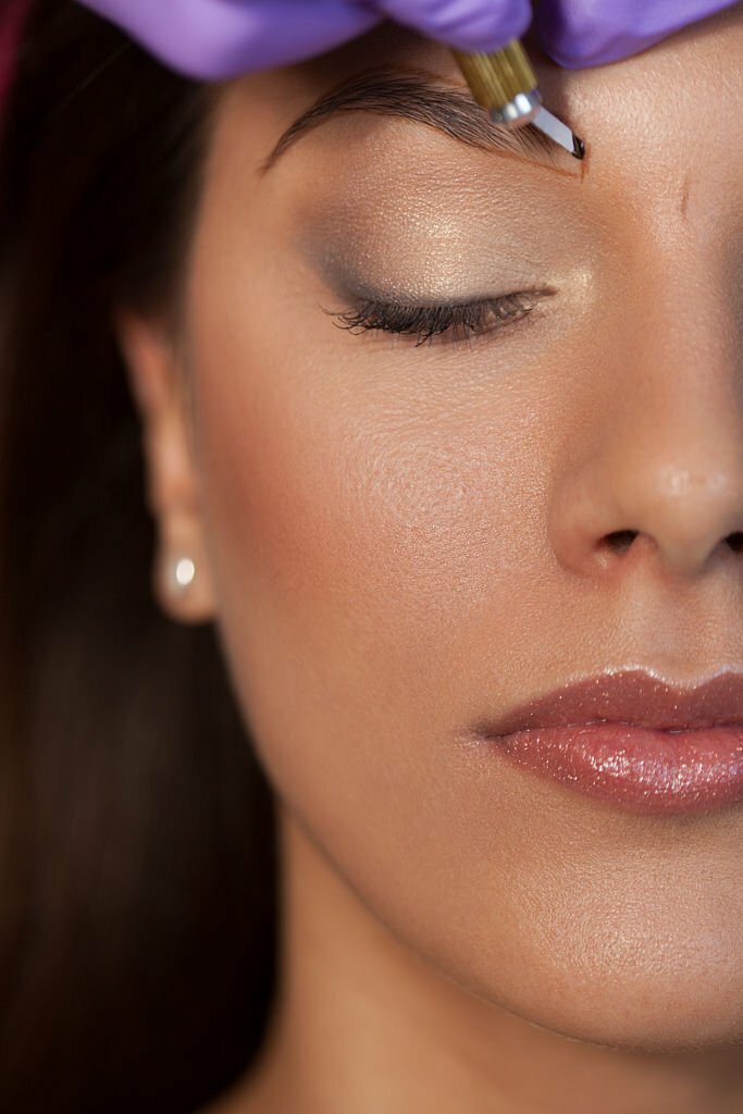 Beautiful young woman is in a beauty salon on the treatment for permanent eyebrow makeup. Woman lying with closed eyes while beautician appling permanent make up with tattoo tool on hers eyebrow. Before that beautician did a the precise shaping eyebrows with crayon. Beautician has purple gloves on hands. Close up shot